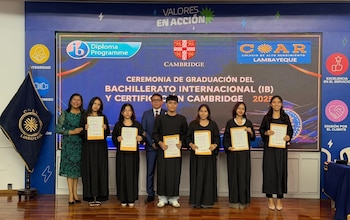 Un grupo de seis estudiantes con togas negras y dos adultos posan sonrientes en un escenario frente a una pantalla con logos de Cambridge y COAR Lambayeque, mostrando sus certificados
