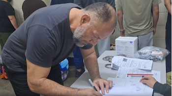 Un hombre con barba y camiseta oscura firma un cuaderno sobre una mesa. En la mesa hay documentos, esposas metálicas, una caja blanca y una bolsa transparente. Otros hombres en el fondo