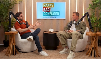 Dos hombres sentados en sillones, con micrófonos de brazo, mesas de madera y plantas. Un monitor con el logo 'Happy Sad Confused' se ve al fondo