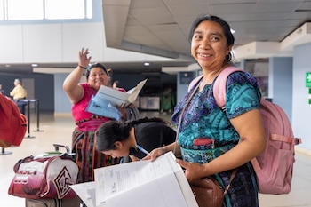 Mujer sonriente con vestimenta tradicional, mochila rosa y documentos, junto a otras personas que rellenan papeles, maletas y una cartera