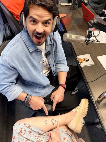 Hombre con barba, auriculares y camisa vaquera azul mira a cámara con boca abierta mientras señala un tatuaje temporal en una pierna junto a un micrófono de estudio