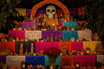 Altar escalonado decorado con papel picado multicolor, calaveras, esqueletos, velas encendidas, fotos antiguas y un arco de flores anaranjadas.