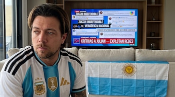 Un hombre con camiseta de Argentina mira a una pantalla de televisión que muestra noticias y comentarios de redes sociales. Una bandera de Argentina sobre un sofá.