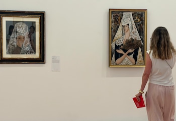 Una mujer de espaldas con un libro rojo observa dos cuadros enmarcados colgados en una pared blanca de una galería de arte. Hay una tarjeta de información entre los cuadros