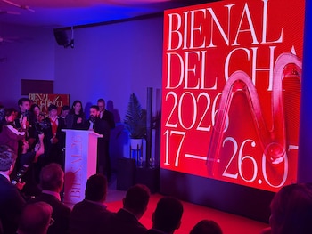 Un orador habla desde un podio blanco con la inscripción 'BIENAL26' frente a un grupo de personas, con una gran pantalla roja que muestra 'BIENAL DEL CHACO 2026' y fechas