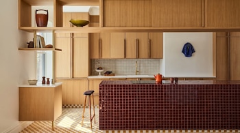 Una cocina moderna presenta gabinetes y estantes de madera clara, una isla central de baldosas rojas, un taburete y un suelo de cuadros amarillos y blancos.