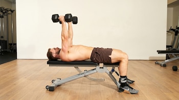 Hombre caucásico sin camiseta, acostado en un banco de pesas gris, sosteniendo dos mancuernas negras con los brazos extendidos hacia arriba en un gimnasio.