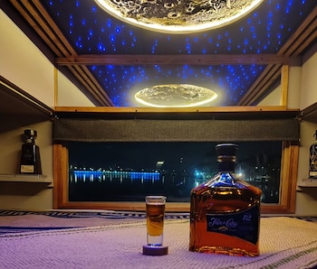 Botella de ron Flor de Caña 12 y vaso de shot con líquido ámbar sobre mesa. Ventana con vista nocturna a lago iluminado. Techo con luces y objeto circular