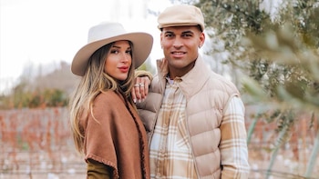 Agustina Gandolfo con sombrero crema y poncho marrón, junto a Lautaro Martínez con gorra plana y chaleco beige, posan sonriendo al aire libre