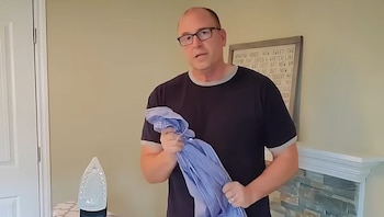 Hombre con anteojos y camiseta oscura sostiene una camisa azul arrugada junto a una plancha
