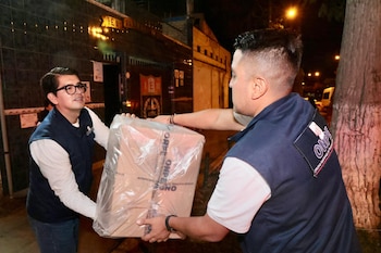 Dos hombres en chalecos ONPE intercambian una caja grande de cartón, envuelta en plástico, en un entorno exterior nocturno junto a un árbol