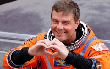 Primer plano del astronauta Reid Wiseman con un traje espacial naranja, sonriendo y haciendo un gesto de corazón con las manos. Se ve su placa con el nombre y la bandera de EE. UU