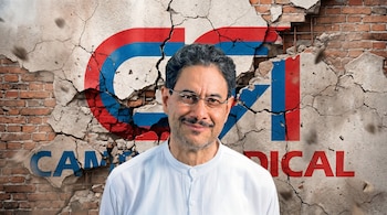 Primer plano de un hombre sonriente con camisa blanca y gafas frente a un muro de ladrillos parcialmente cubierto por yeso agrietado con el logo de 'CAMBIO RADICAL'.