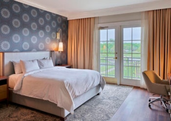 Habitación de hotel con cama king-size blanca, mesitas de noche, lámpara de pared y silla de escritorio. Ventana con puertas francesas y balcón exterior visible