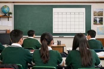 Vista trasera de estudiantes de secundaria con uniforme verde sentados en un salón de clases mexicano. Al fondo, un pizarrón con un calendario de abril y un globo terráqueo.