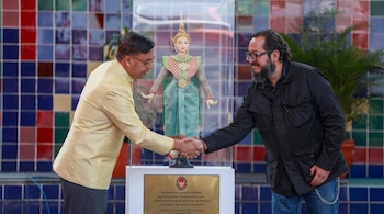 Dos hombres estrechan manos frente a una vitrina transparente. Dentro, una muñeca con atuendo tradicional tailandés. Una placa dorada está debajo de la vitrina.