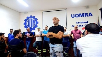 Angel Derosso, un hombre calvo con camiseta negra y jeans, de pie con los brazos cruzados, habla ante un grupo de hombres en una sala con el logo de UOMRA