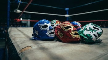 Cinco máscaras de lucha libre de colores azul, rojo, verde y blanco están colocadas en el suelo de un ring con cuerdas rojas y blancas.