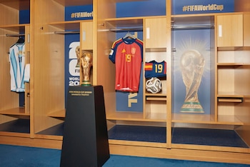 Vestuario con camisetas de Argentina y España, trofeo Copa Mundial FIFA en pedestal, balón de fútbol y pósters del trofeo en taquillas de madera