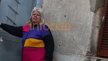 Mujer de cabello rubio, con suéter colorido (negro, rosa, morado, amarillo), rostro entristecido, de pie junto a una pared de hormigón desgastada y una persiana de madera
