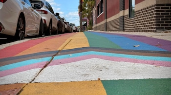 Acera de San Antonio pintada con los colores de la bandera Progress Pride, vista desde abajo, con coches aparcados y edificios a los lados bajo un cielo azul