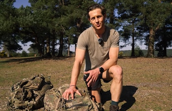 Hombre arrodillado en un terreno boscoso, con camiseta caqui y pantalones cortos, junto a dos mochilas de camuflaje. Al fondo, un bosque de pinos bajo cielo azul