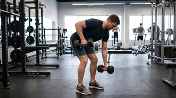 Un hombre inclinado sostiene dos mancuernas en un gimnasio con soportes de pesas, bancos y máquinas de ejercicio.