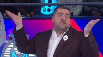 Un hombre con barba, traje marrón, camisa blanca y un adorno floral eleva las manos en un plató iluminado con luces de colores.