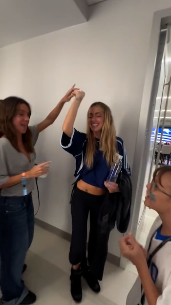 Tres personas. Una mujer rubia con camiseta de fútbol levanta un brazo y sonríe, sosteniendo una botella. Una mujer de cabello castaño levanta un brazo. Una persona con la cara pintada de azul mira a la mujer rubia