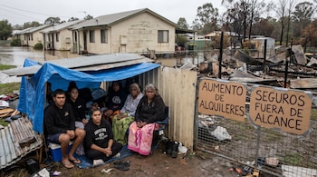 Personas indígenas australianas bajo un refugio improvisado. Al fondo, casas inundadas y destrozadas por incendios. Letreros sobre alquileres y seguros inalcanzables.