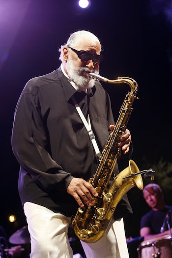Sonny Rollins, con barba blanca y gafas de sol, toca el saxofón tenor en un escenario oscuro, vistiendo una camisa negra y pantalones blancos