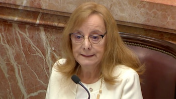 Primer plano de Alicia Kirchner, mujer de pelo rubio y gafas, con una chaqueta blanca, sentada frente a un micrófono con un fondo de mármol rojizo