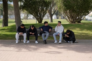 Cinco jóvenes con ropa deportiva en un banco de madera y en el suelo de un parque arbolado junto a un camino pavimentado.