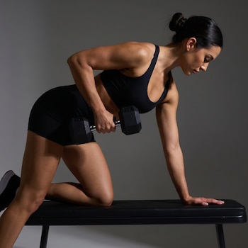 Vista lateral de una mujer en ropa deportiva negra, apoyada en un banco y levantando una mancuerna con su mano derecha.