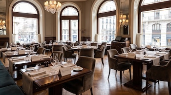 Interior de un restaurante con mesas de madera, sillas, mantelería y cubiertos. Cada mesa tiene un cartel de “Reservado”. Ventanales altos, arañas y espejos.