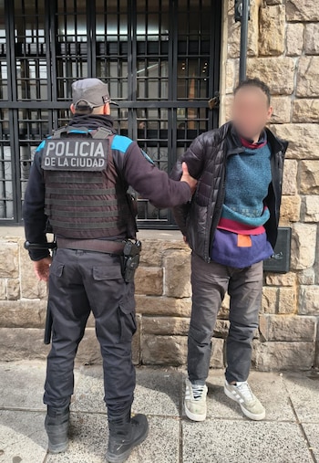 Un agente de la Policía de la Ciudad de espalda sujeta a un hombre detenido con el rostro difuminado junto a un muro de piedra