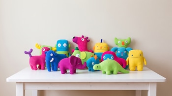 Un grupo de doce peluches de colores brillantes sobre una mesa blanca, incluyendo formas de elefantes, un dinosaurio y figuras abstractas.