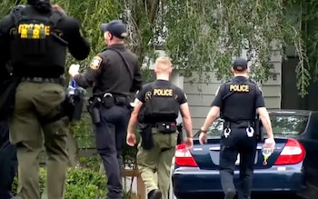 Cuatro agentes de policía con chalecos negros caminan de espaldas hacia una vivienda cerca de un automóvil azul