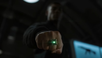 Un puño cerrado en primer plano con un anillo que emite luz verde. Una persona desenfocada en el fondo oscuro con un objeto azul brillante