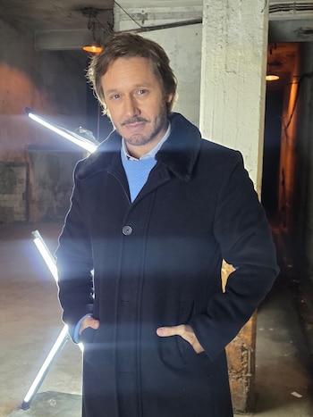 Hombre con barba y bigote, vestido con abrigo oscuro, suéter azul claro y camisa blanca, posa con las manos en los bolsillos en un espacio oscuro con luces