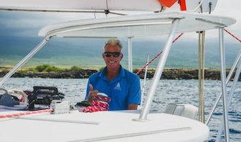 Hombre de mediana edad con gafas de sol y camisa azul sonríe y hace un pulgar hacia arriba desde el asiento del capitán de un velero, con el mar y la costa rocosa de fondo