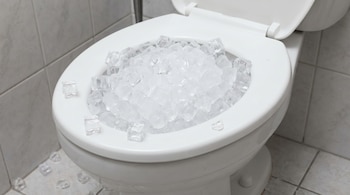 Un inodoro blanco con la tapa levantada, completamente lleno de cubos de hielo transparentes. Algunos cubos de hielo están esparcidos en el suelo de baldosas grises.