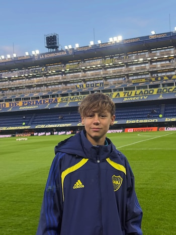 Niño rubio vestido con chaqueta azul y amarilla de Boca Juniors, de pie sobre el césped de un estadio de fútbol, con las gradas de La Bombonera al fondo