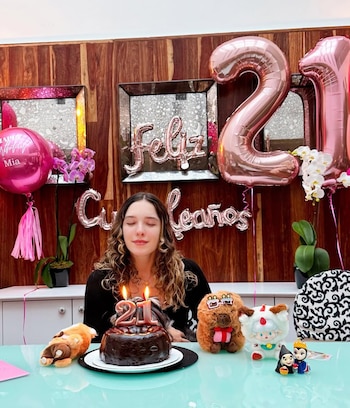 Mujer joven con ojos cerrados frente a pastel de chocolate con velas del número 21. Globos de 'Feliz Cumpleaños' y '21' en rosa decoran el fondo