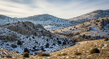 Montañas rocosas en Grecia con parches dispersos de nieve, intercalados con vegetación seca y arbustos bajos bajo un cielo claro con nubes finas.