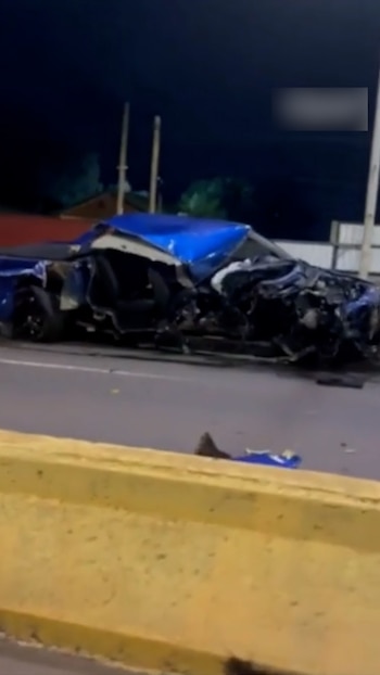 Camioneta azul oscuro destrozada en una carretera por la noche, con el frontal muy dañado y el motor visible, junto a una barrera amarilla