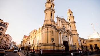 Catedral amarilla con dos torres, una gran cruz en la pared exterior, rejas de hierro, escalones con personas y automóviles en la calle.