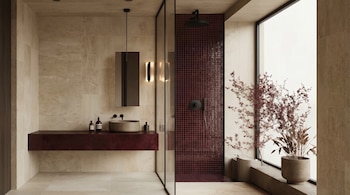 Un baño moderno con paredes de azulejos beige, un mueble bajo granate con lavabo redondo, una ducha con azulejos pequeños rojos y una ventana grande con una planta.