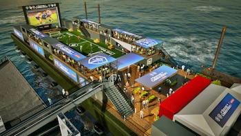 Vista aérea de una plataforma flotante para un evento de fútbol, con césped artificial, pantallas de video, áreas de asientos y logos de celebración sobre el agua