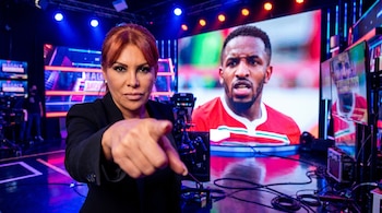 Magaly Medina en primer plano con cabello rojo y blazer negro, apuntando a la cámara. Detrás, en una pantalla LED, se ve a Jefferson Farfán en un set de TV.